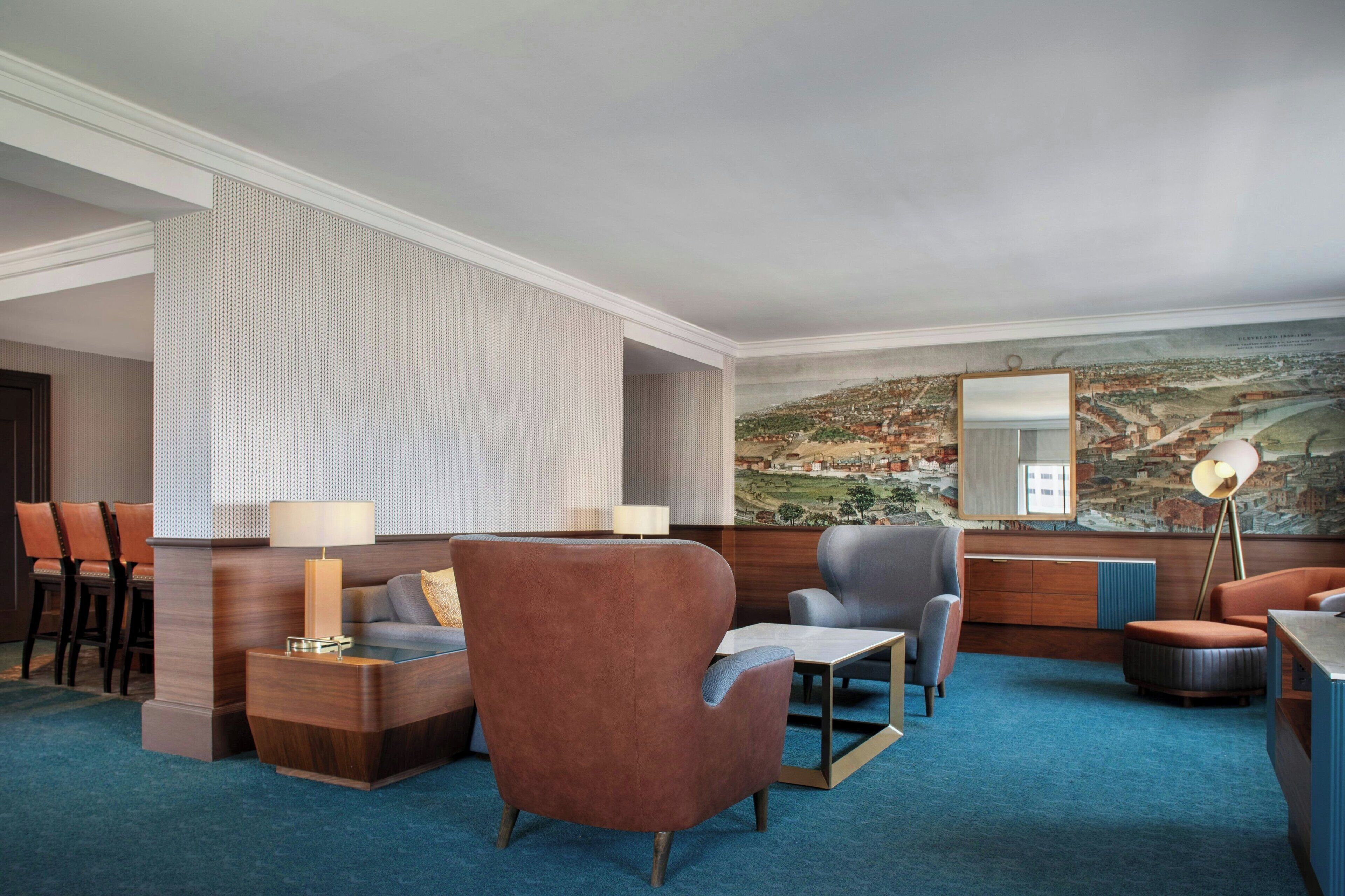 A hotel lobby with a blue carpet, seating area with sofas and chairs, a dining table, a lamp, and a large mural on the wall.