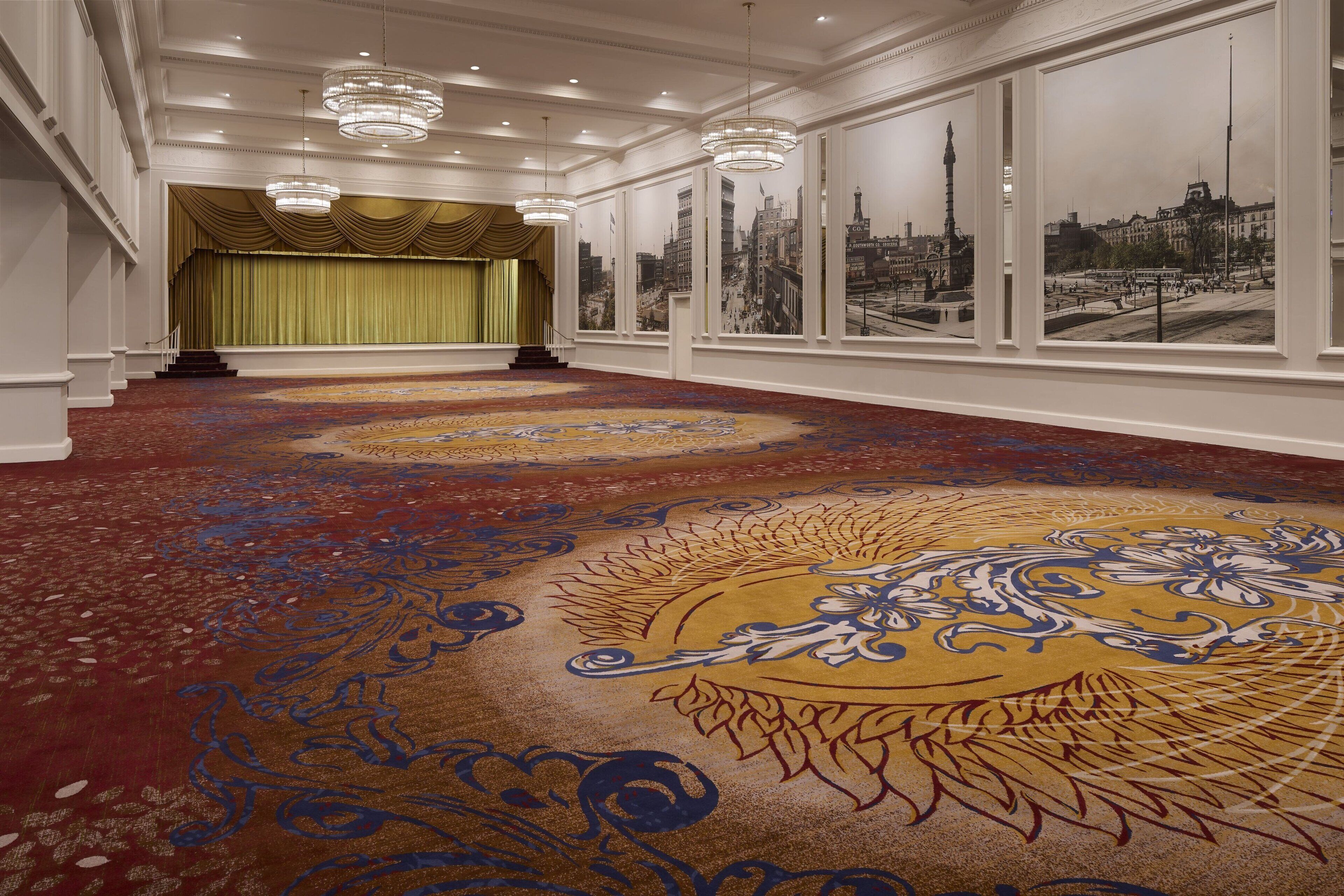 A spacious hallway with a patterned carpet, large windows with cityscape views, and a stage with a green curtain.