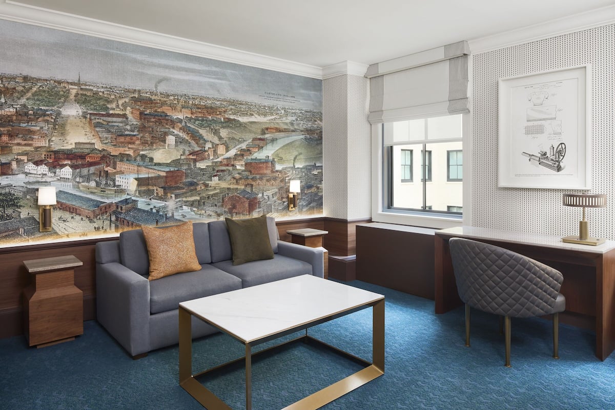 A modern hotel room with a large cityscape mural, a comfortable sofa, a desk with a chair, and a small side table.