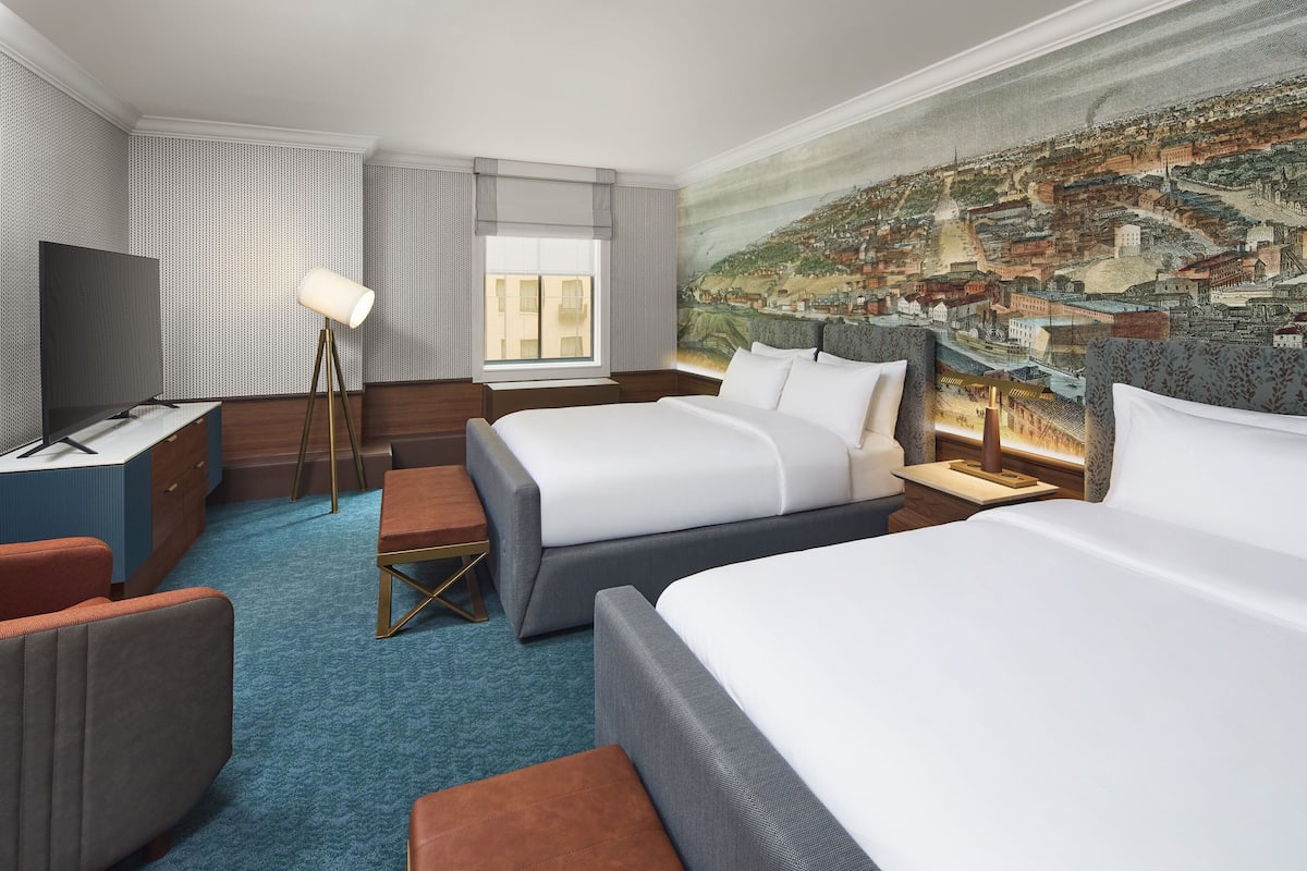 A hotel room with two beds, a desk with a TV, a lamp, and a large cityscape mural on the wall.