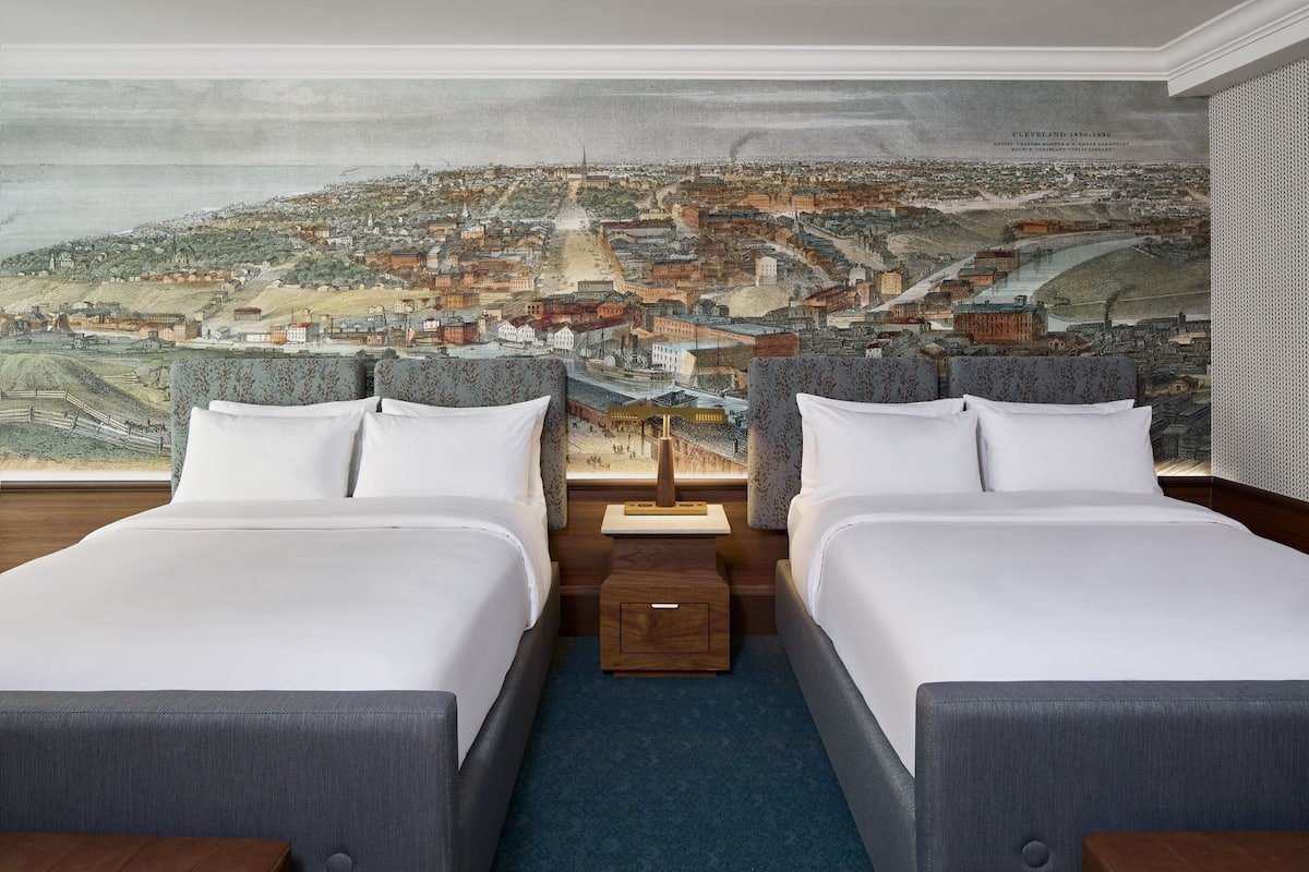 A hotel room with two beds, a large cityscape mural on the wall, and a small wooden nightstand between the beds.