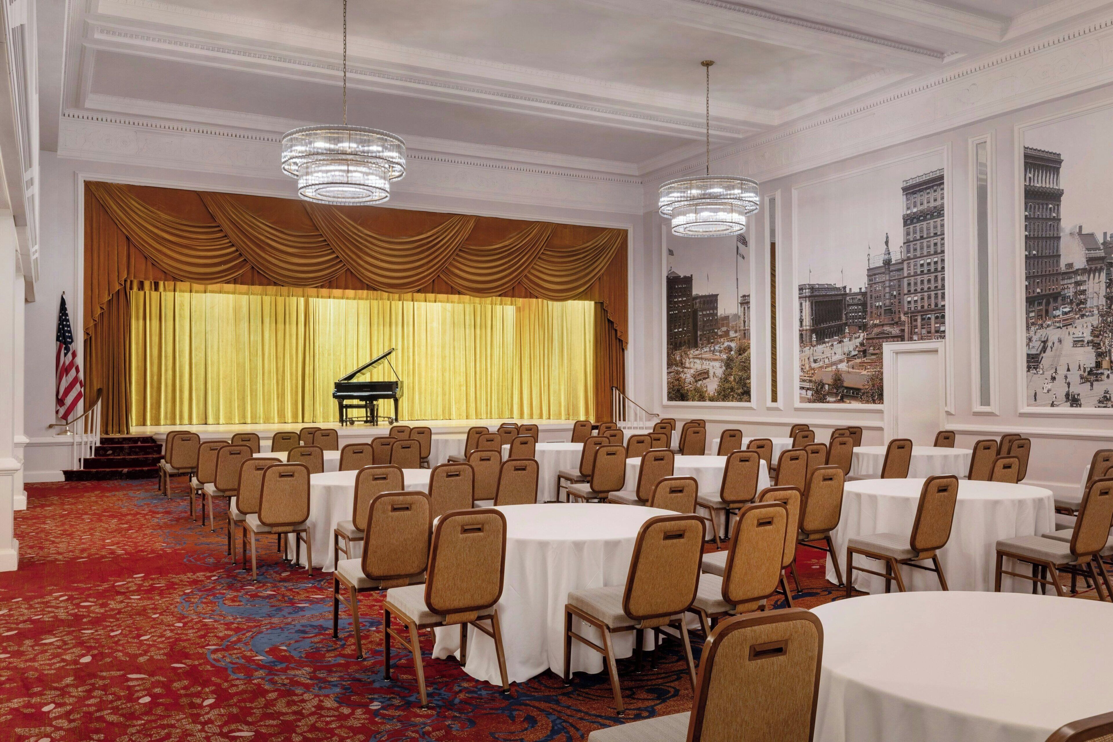 A conference room with rows of tables and chairs, a stage with a piano, and large windows with city views.