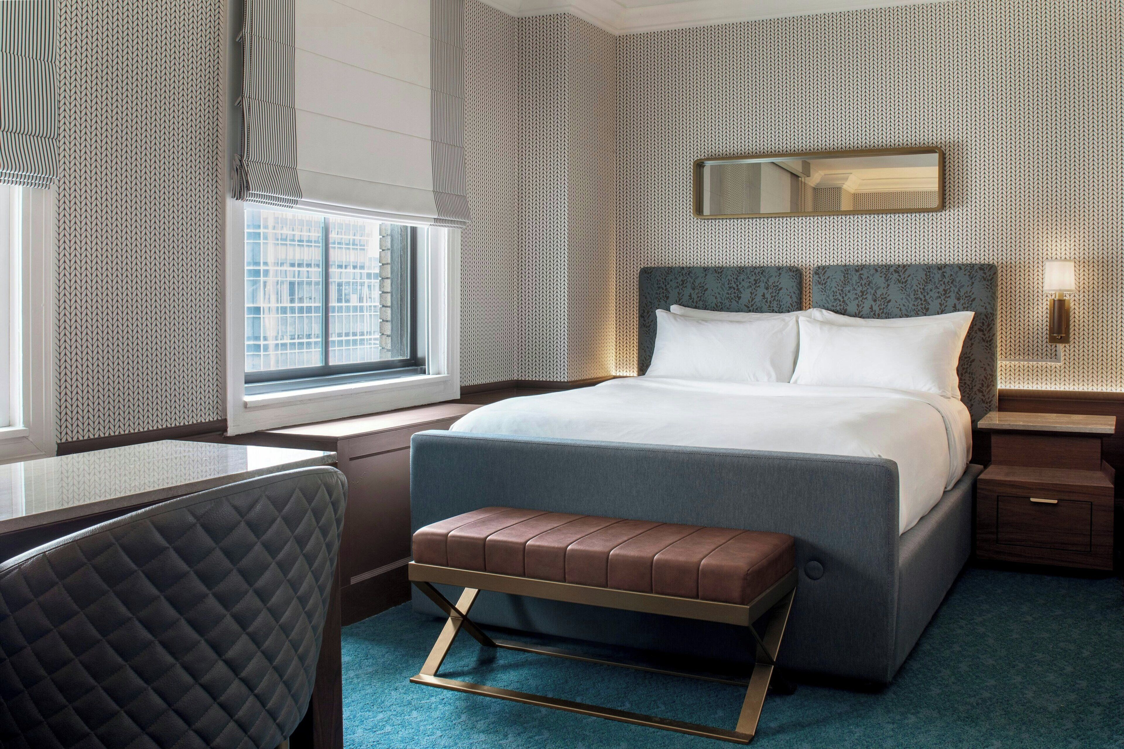 Habitación de hotel con cama, escritorio, silla, banco, ventana y espejo.