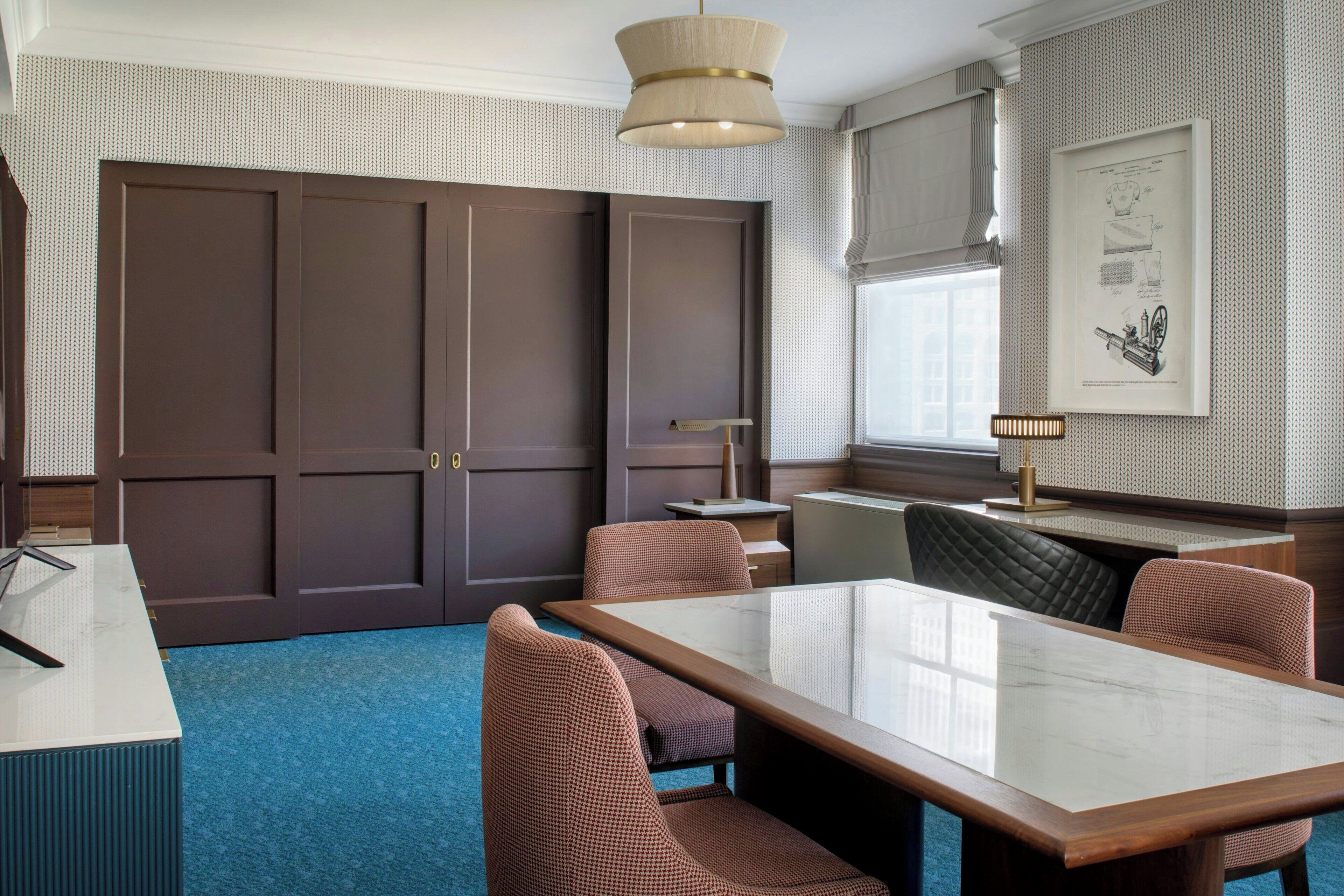 A hotel room with a large wardrobe, a dining table with chairs, a desk, a lamp, and a window with a view.