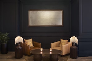 A dark room with a large framed artwork, two leather sofas with patterned cushions, a round wooden coffee table, and two side tables with lamps and candles.