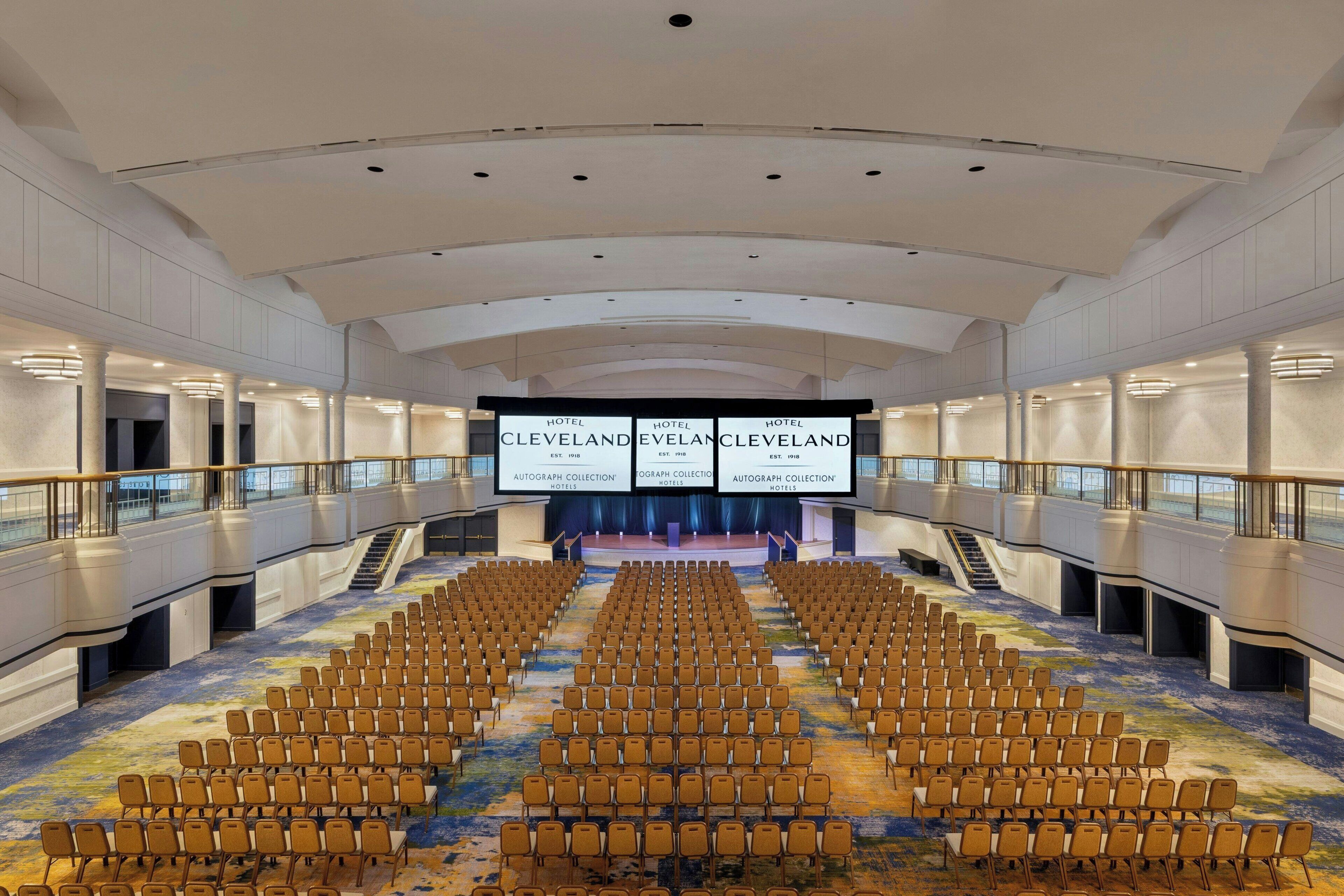 A large, modern hotel auditorium with rows of chairs, a stage, and a large screen displaying the hotel's name.