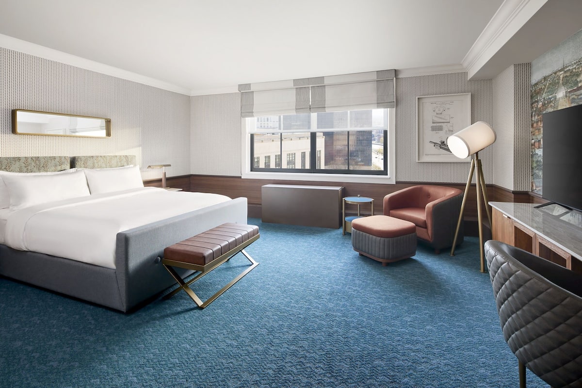 A modern hotel room with a large bed, a desk, a chair, and a view of the city through the window.