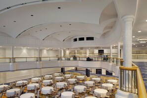 A multi-level hotel lobby with round tables and chairs, a grand staircase, and a high ceiling with recessed lighting.
