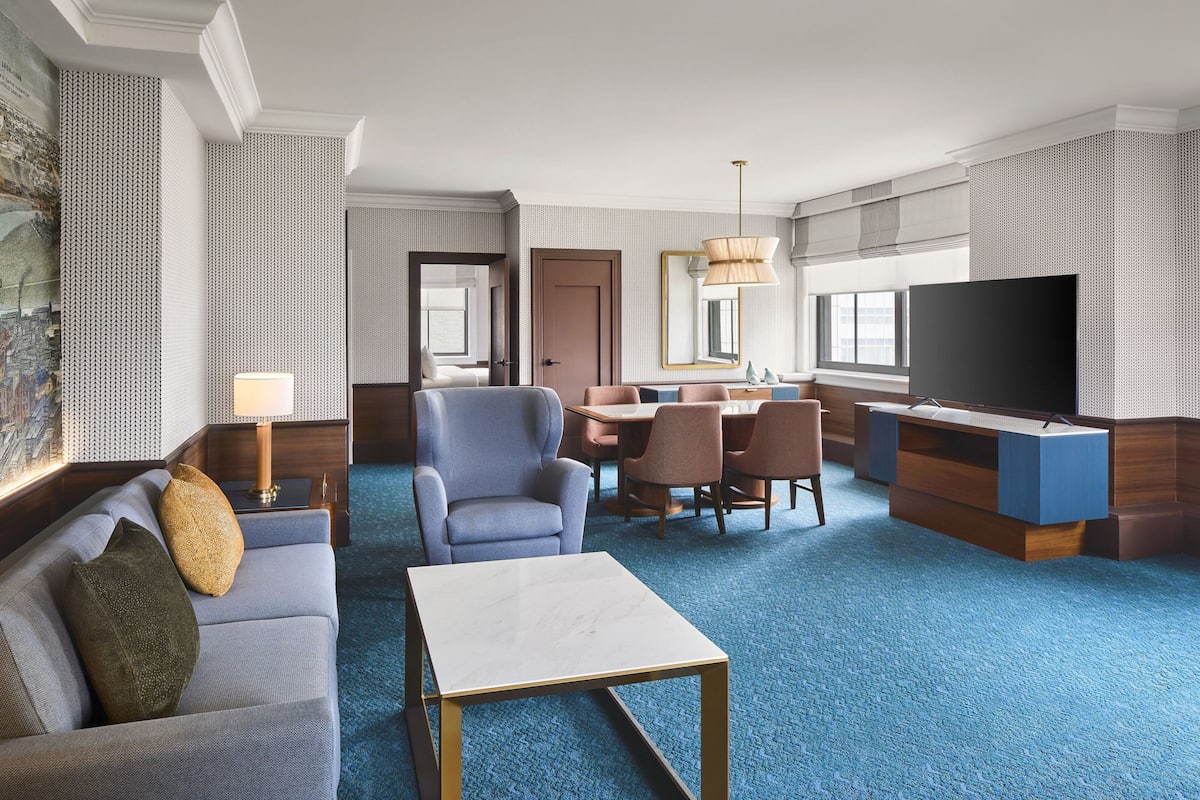 A hotel room with a blue carpet, a sofa, a coffee table, a TV, a dining area with chairs, and a large mirror.