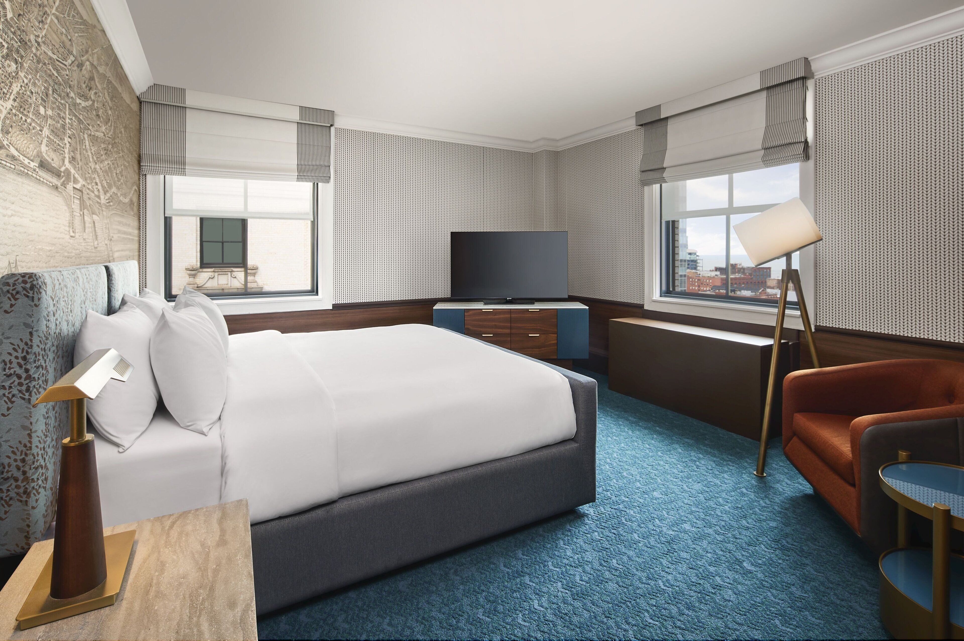A hotel room with a large bed, a flat-screen TV, a desk with a lamp, a chair, and a view of the city through the windows.