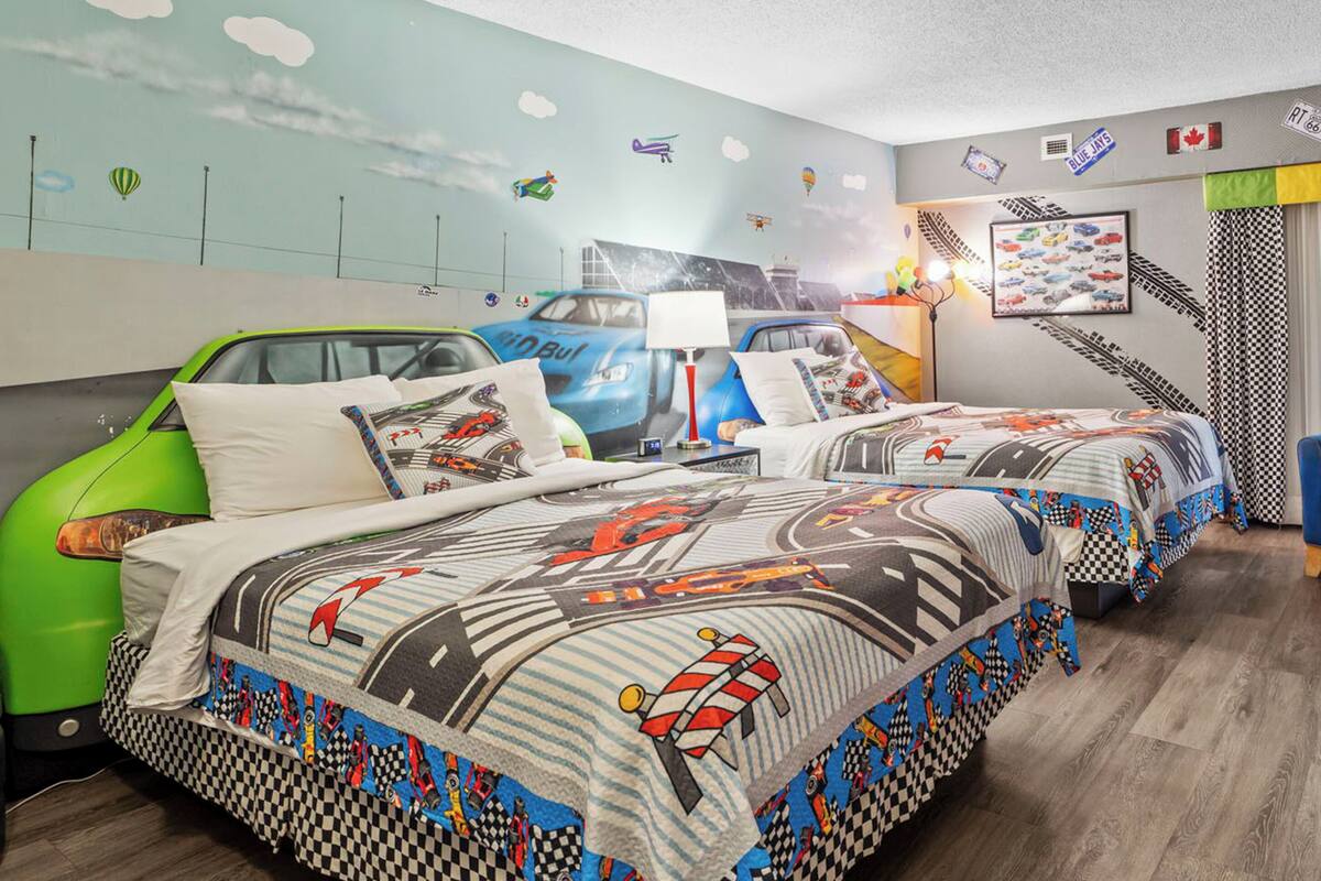 A bedroom with two beds, a wall mural of cars and clouds, a lamp, and a checkered curtain.