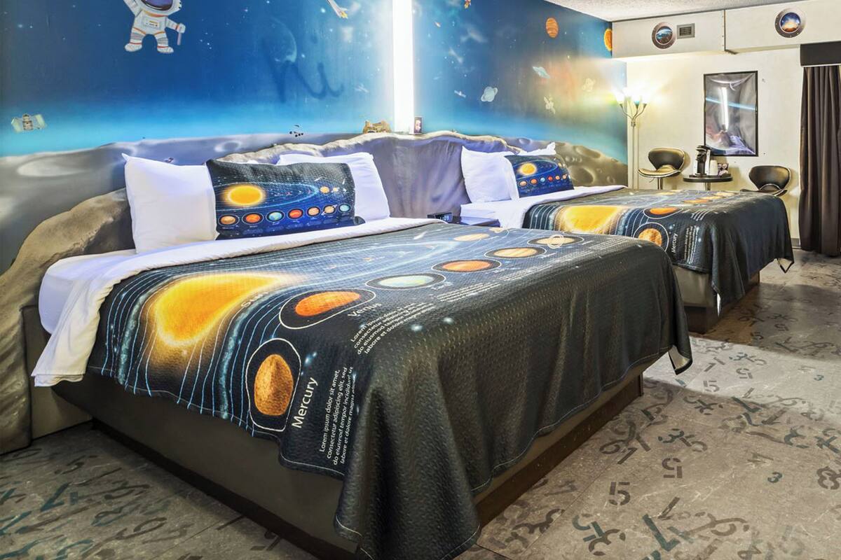 A hotel room with a space-themed bedspread, a mural of an astronaut and planets, and a ceiling light fixture.