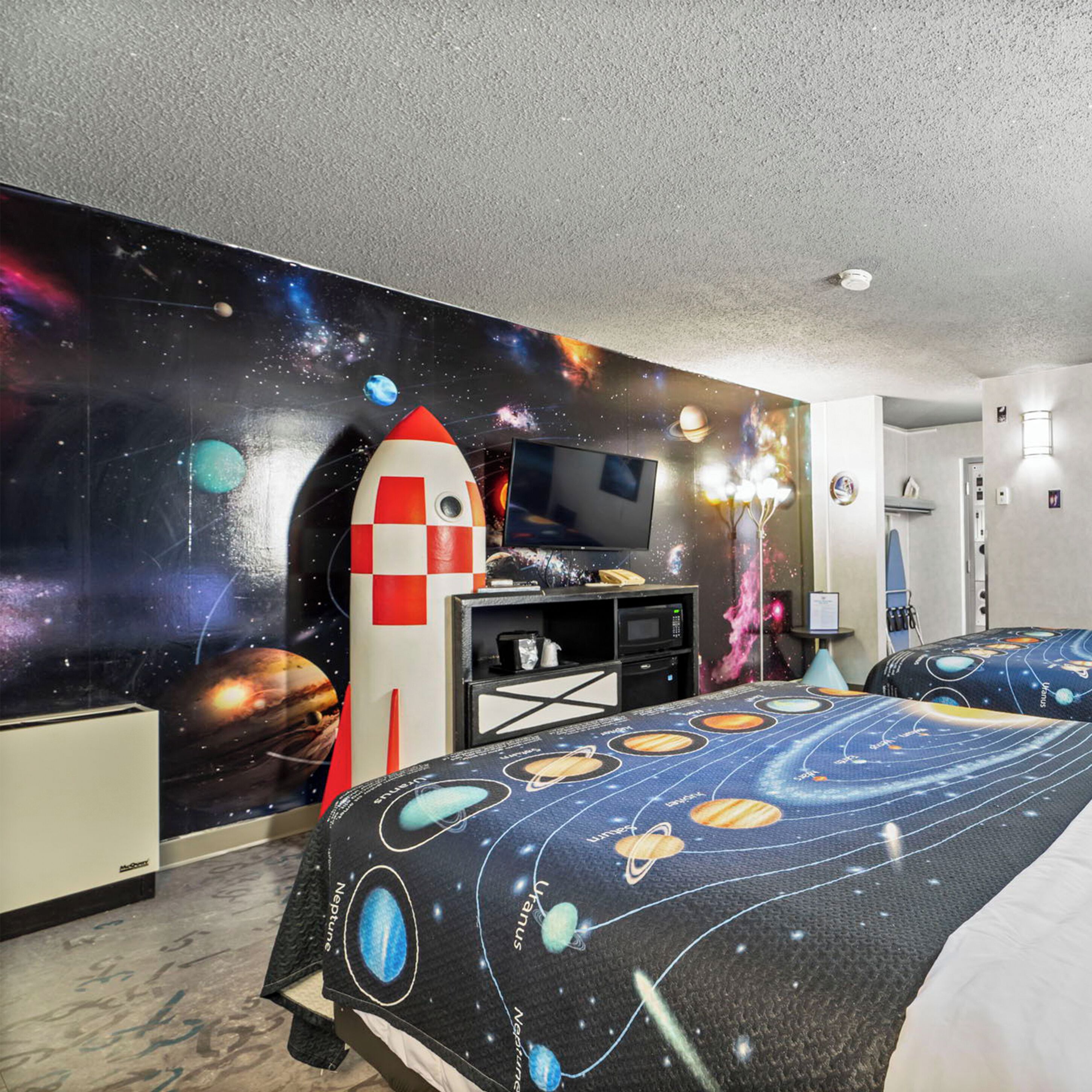 A bedroom with a space-themed wall mural, a bed with a galaxy print bedspread, and a red and white rocket decoration.