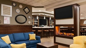 Un espacio lounge moderno con un sofá azul, un sillón amarillo, un televisor de pantalla plana en la pared, una zona de bar con taburetes y una chimenea.