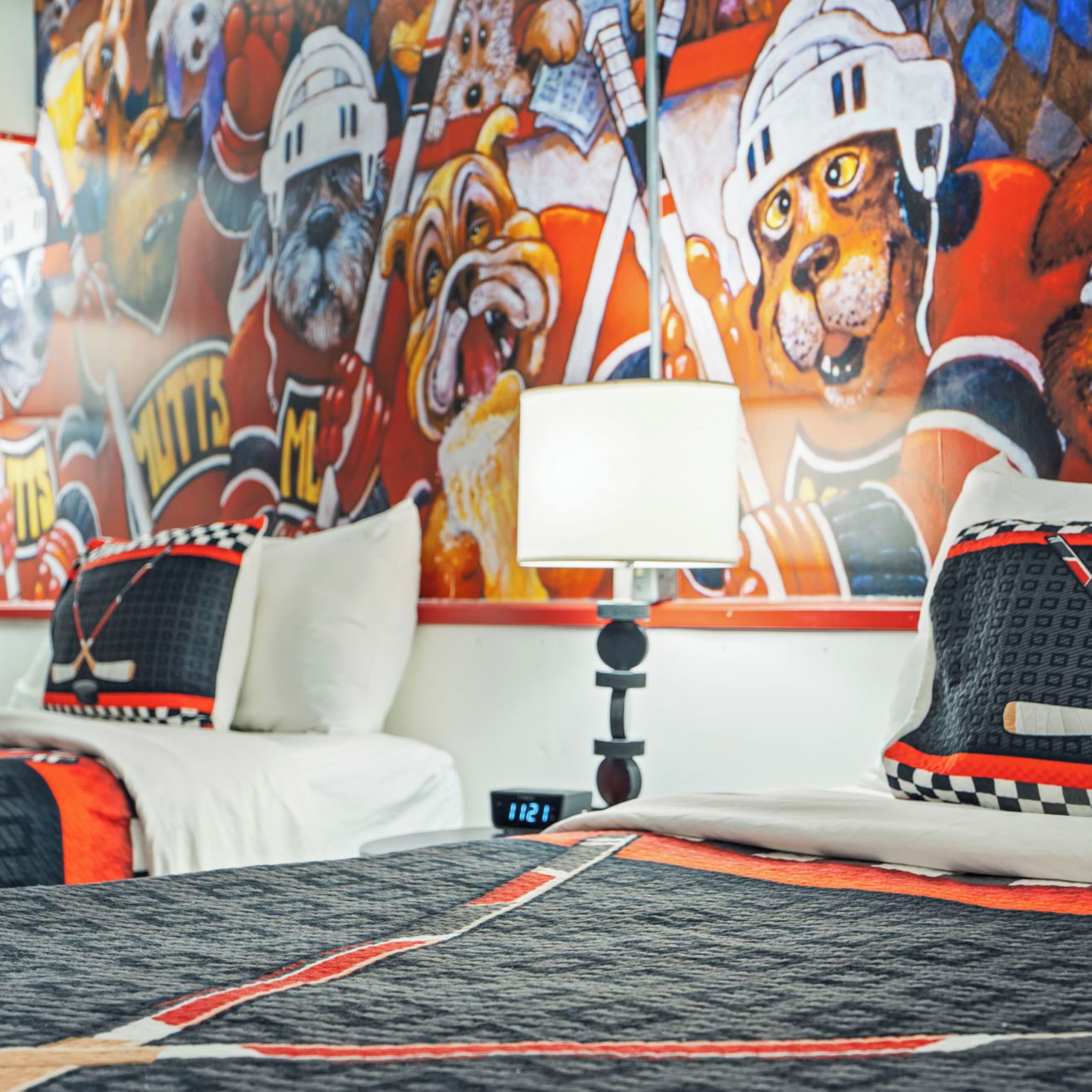 A hotel room with a large sports-themed mural on the wall, a bed with a hockey jersey pillow, and a digital clock on the nightstand.