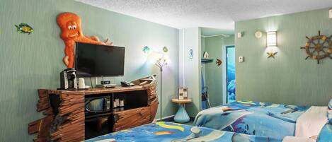 A room with a bed featuring a marine-themed bedspread, a wooden dresser with a television, and a wall-mounted light fixture.