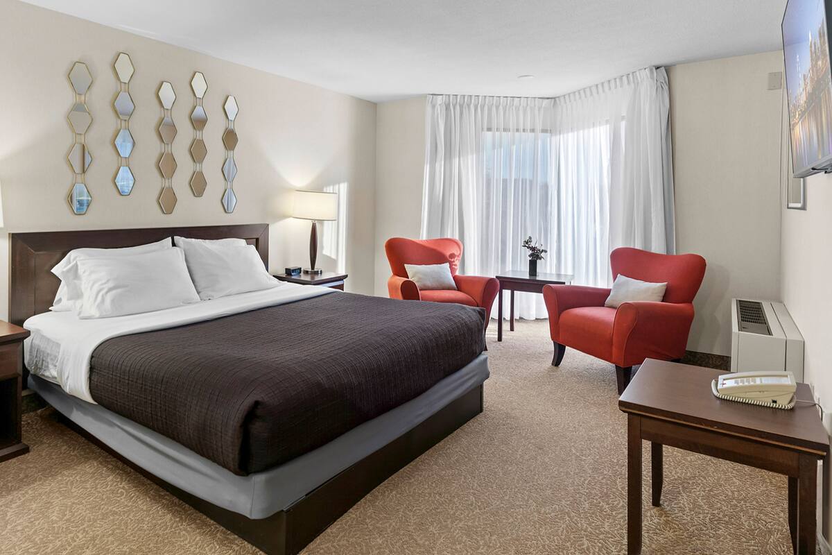 A hotel room with a large bed, two red armchairs, a small table, a TV, and a wall-mounted mirror.