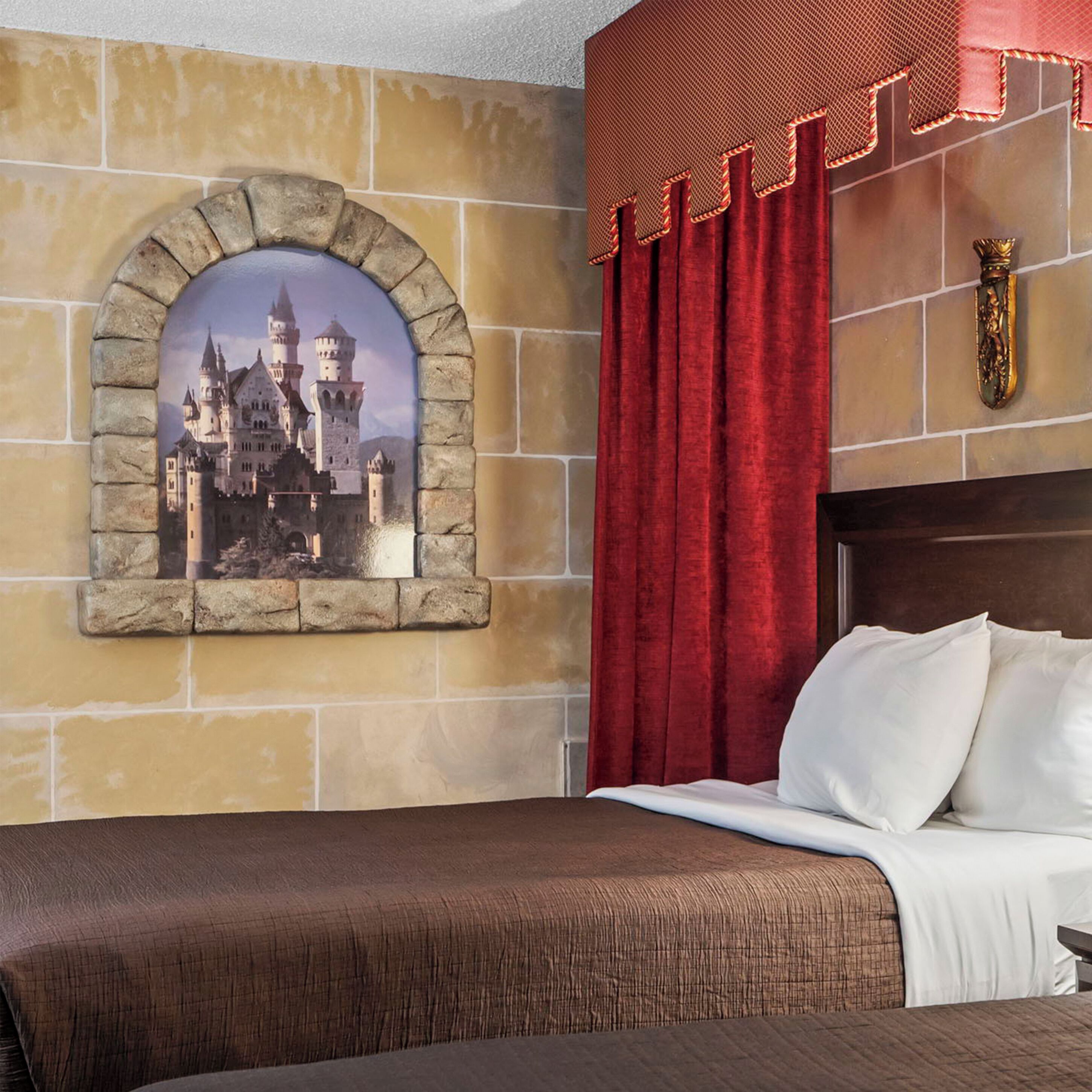 Un dormitorio con un cuadro de castillo en marco de piedra, cortinas rojas y una cama con almohadas blancas.