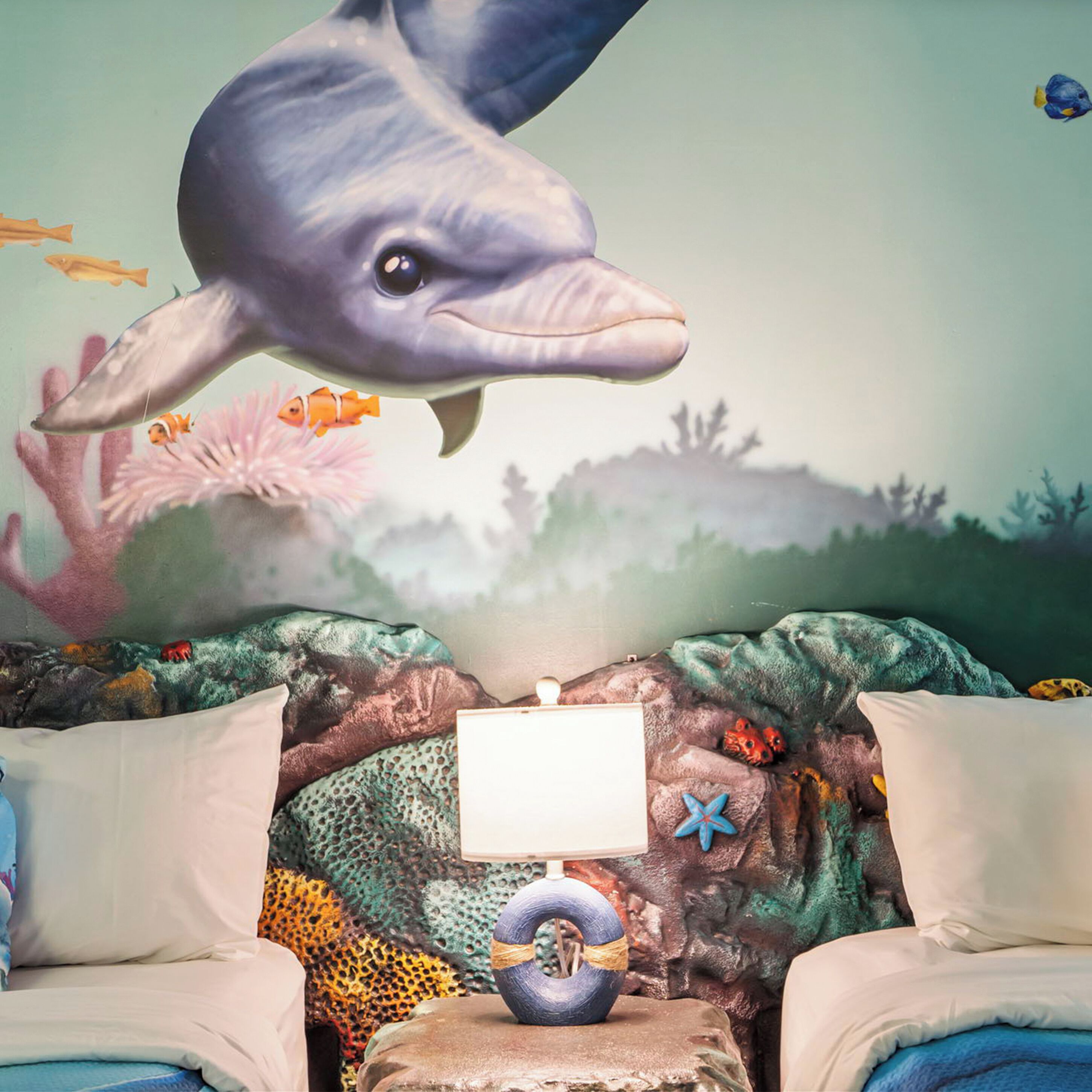 Un dormitorio con un mural de un delfín, una cama, almohadas y una mesita de noche con una lámpara.