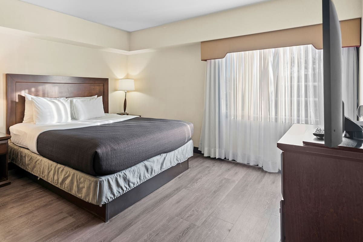 A hotel room with a large bed, a bedside table with a lamp, a desk with a computer, and a window with curtains.