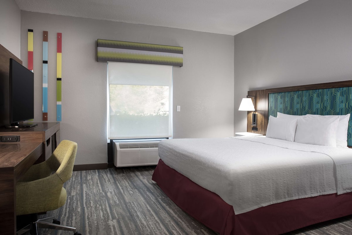 A hotel room with a bed, a desk with a chair, a TV, a window, and a wall-mounted air conditioner.