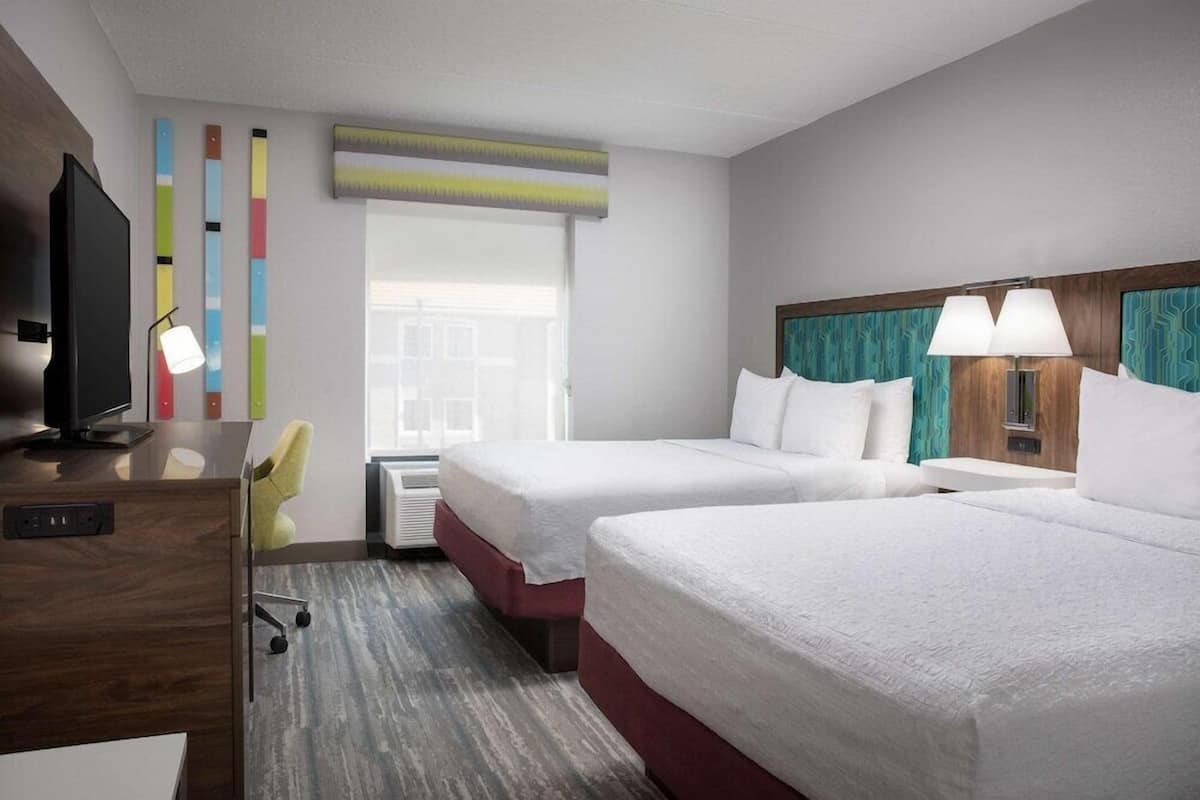 Quarto de hotel com duas camas, uma escrivaninha, uma cadeira, uma televisão e uma janela com persianas.