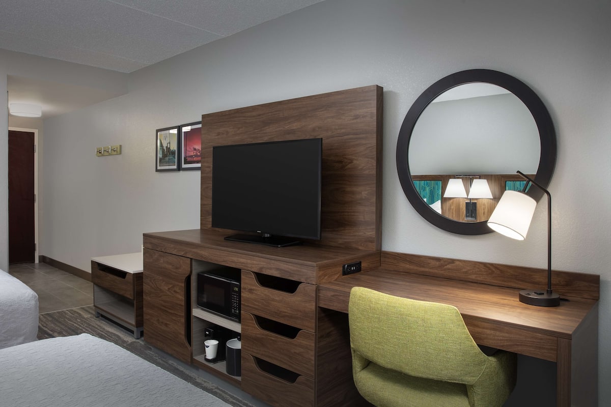 A hotel room with a wooden desk, a flat-screen TV, a mirror, and a green chair.