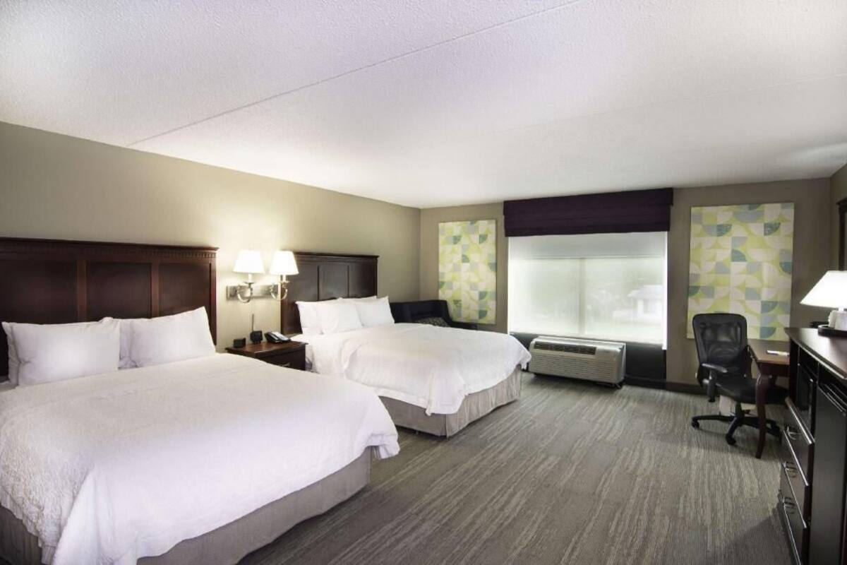 A hotel room with two beds, a desk, a chair, a lamp, a heater, and a window with a view.