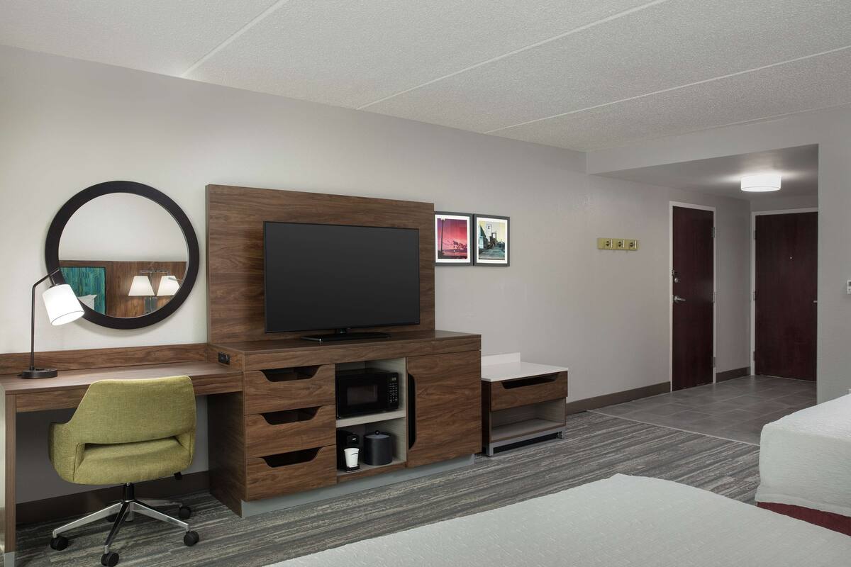 A hotel room with a wooden desk, a television, a green chair, a bed, and a door.