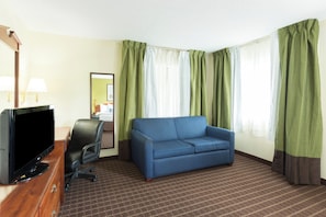 Habitación de hotel con televisor, sofá azul, escritorio con lámpara y cortinas verdes.