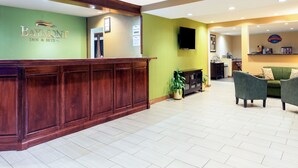 A reception area with a wooden reception desk, a green wall with the hotel name, a flat-screen TV, and a seating area with two chairs and a sofa. - Baymont by Wyndham Louisville South/Shepherdsville/I 65 (Brooks)