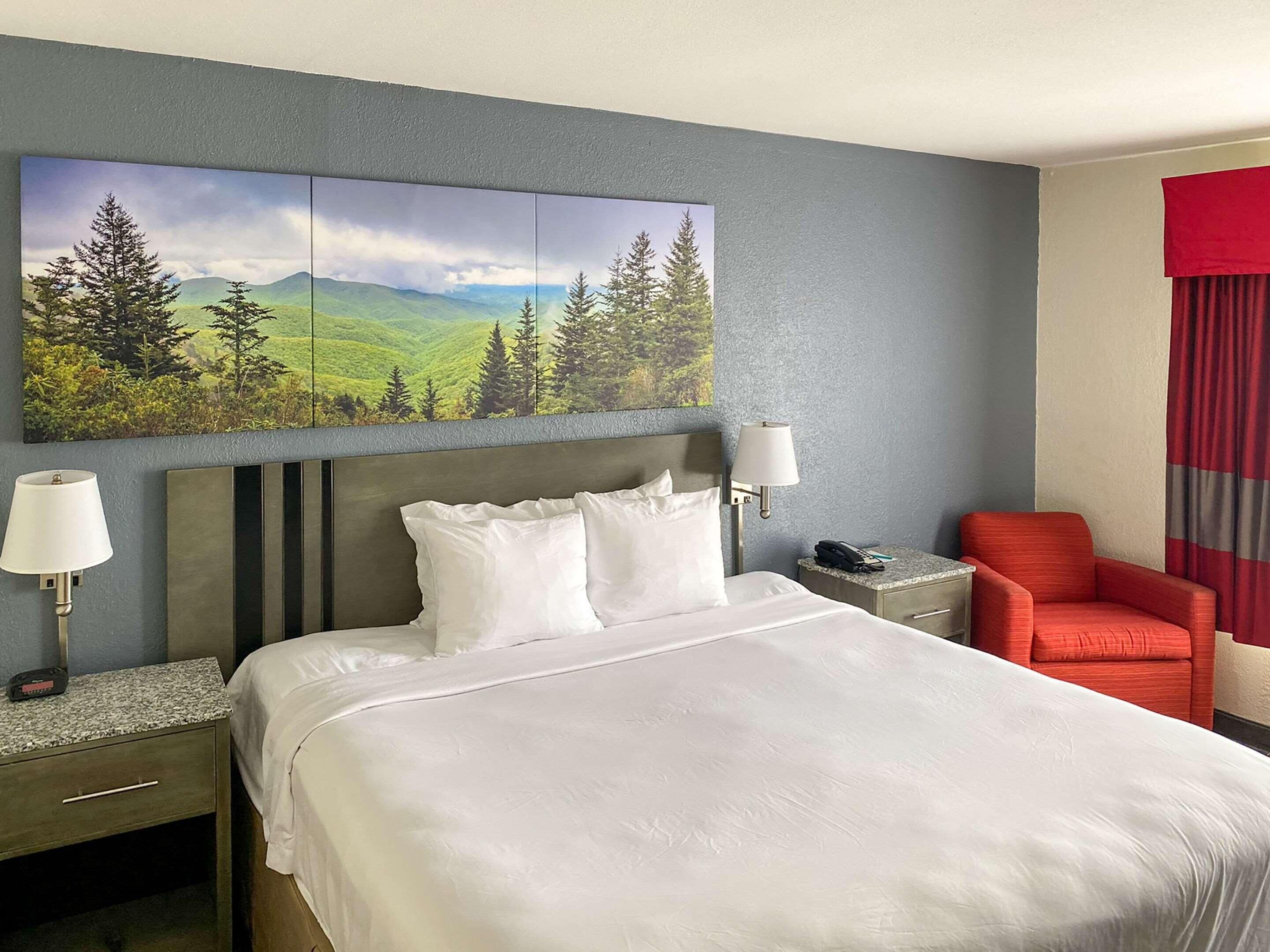 A hotel room with a large bed, two bedside tables, a red armchair, a painting of a landscape, and a red curtain.