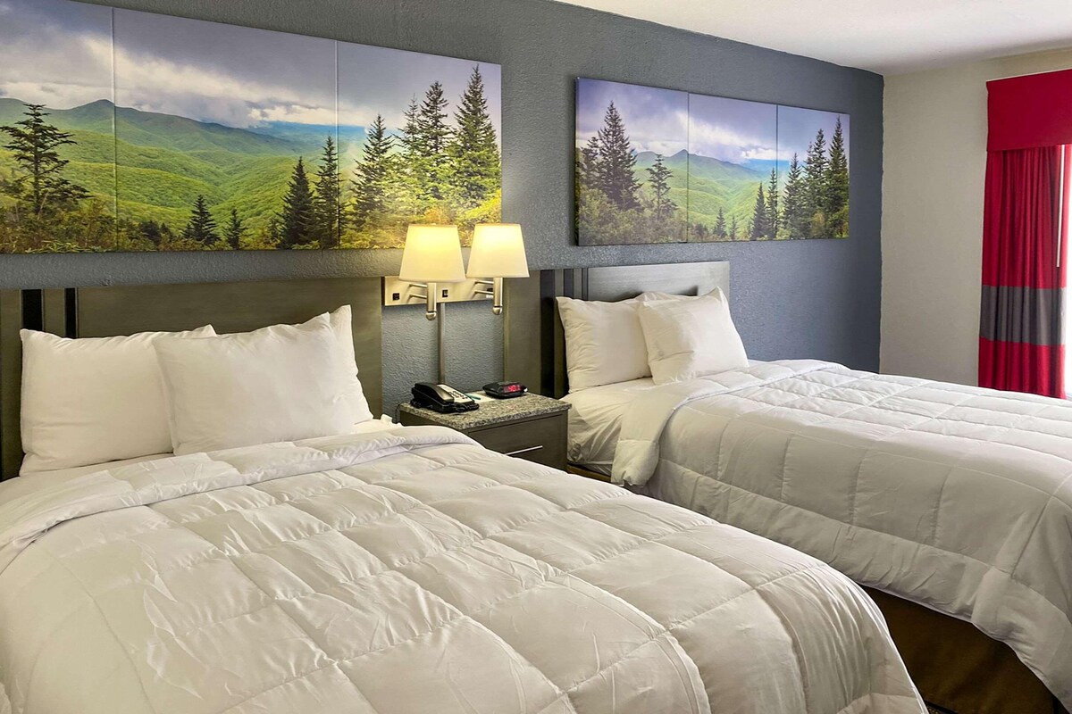 A hotel room with two beds, a nightstand with a phone and lamp, and two wall-mounted paintings of landscapes.