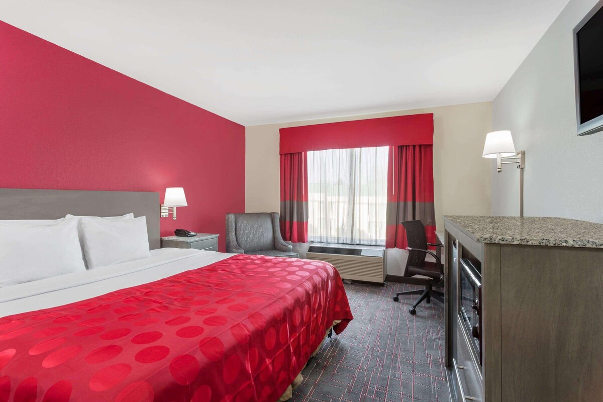 Quarto de hotel com uma cama grande, uma escrivaninha, uma cadeira, uma televisão e uma janela com cortinas.