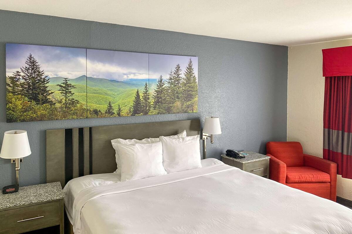 A hotel room with a large bed, two bedside tables, a red armchair, a painting of a landscape, and a red curtain.