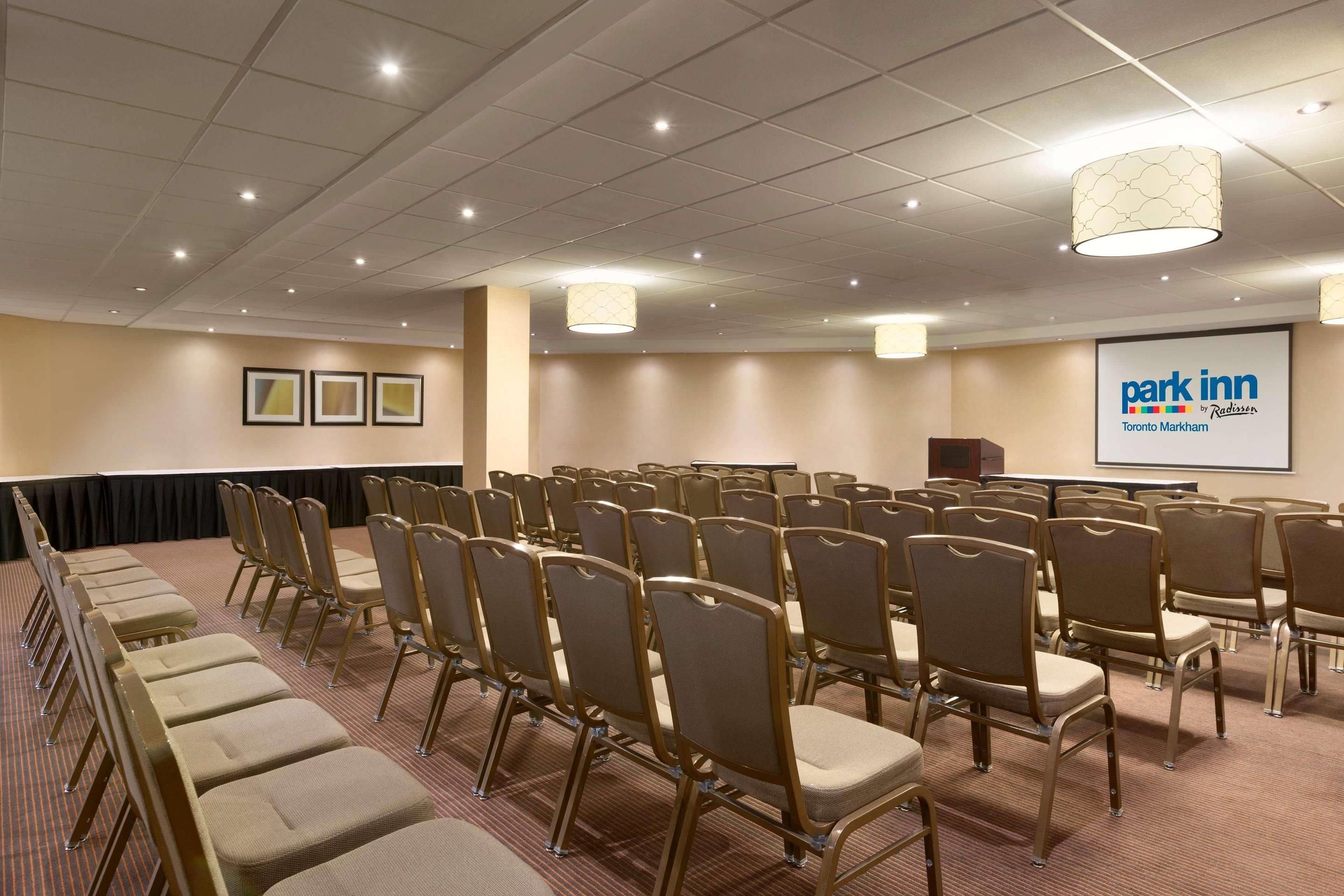 A conference room with rows of chairs, a podium, and a screen displaying 'Park Inn Toronto Markham'.