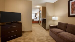 A hotel room with a flat-screen TV on a dark wooden dresser, a brown sofa, a small table with a lamp, and a framed picture on the wall.