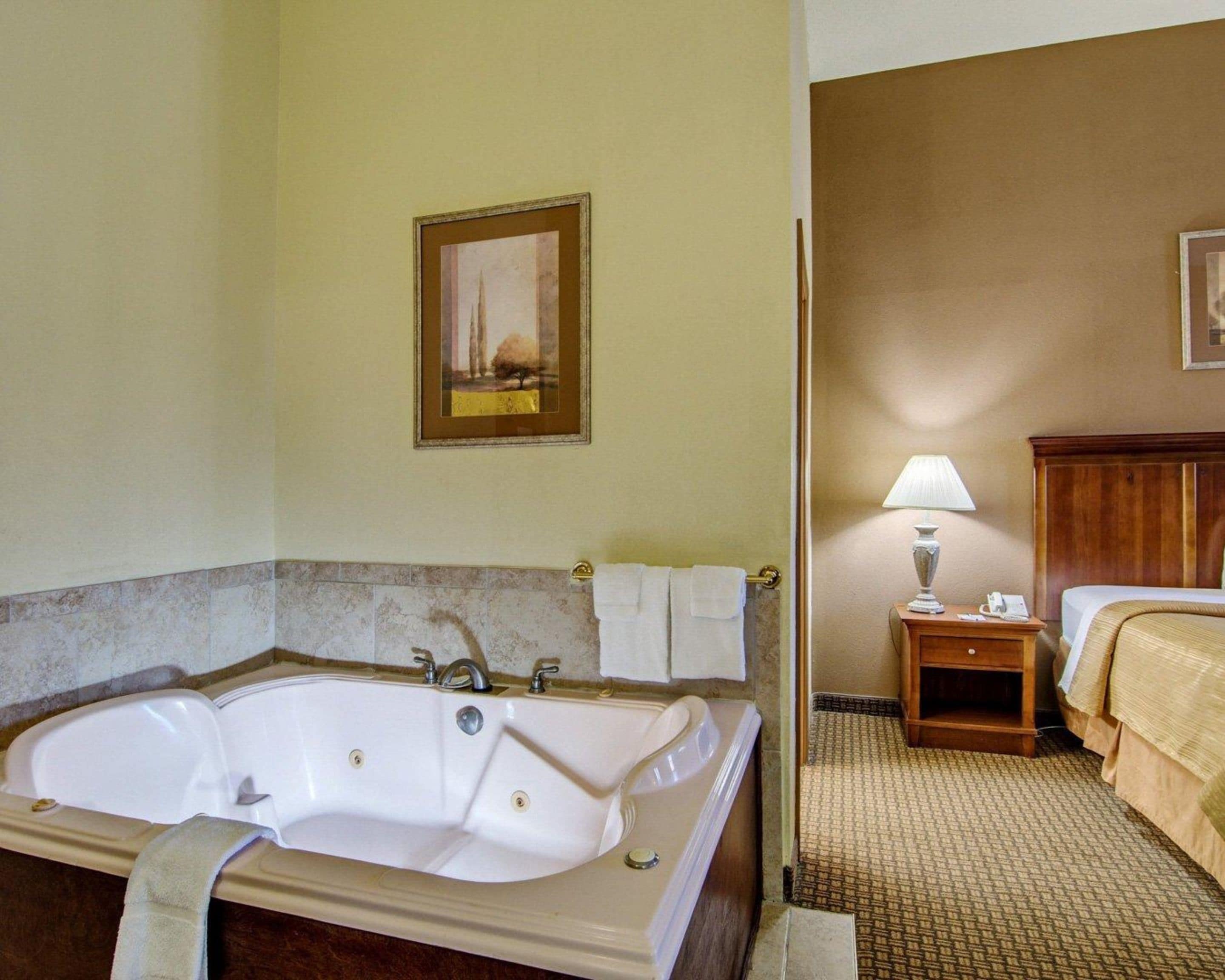A hotel bathroom with a large bathtub, a small bedside table with a lamp, and a bed with a headboard.
