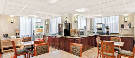A hotel dining area with wooden tables and chairs, a kitchen counter with appliances, and large windows with curtains.