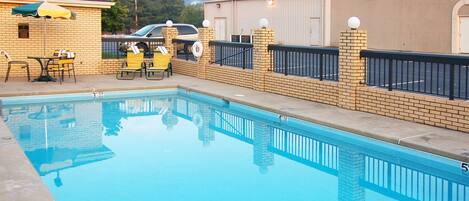 A swimming pool with a brick wall and a fence, a covered seating area with chairs, and a building in the background.