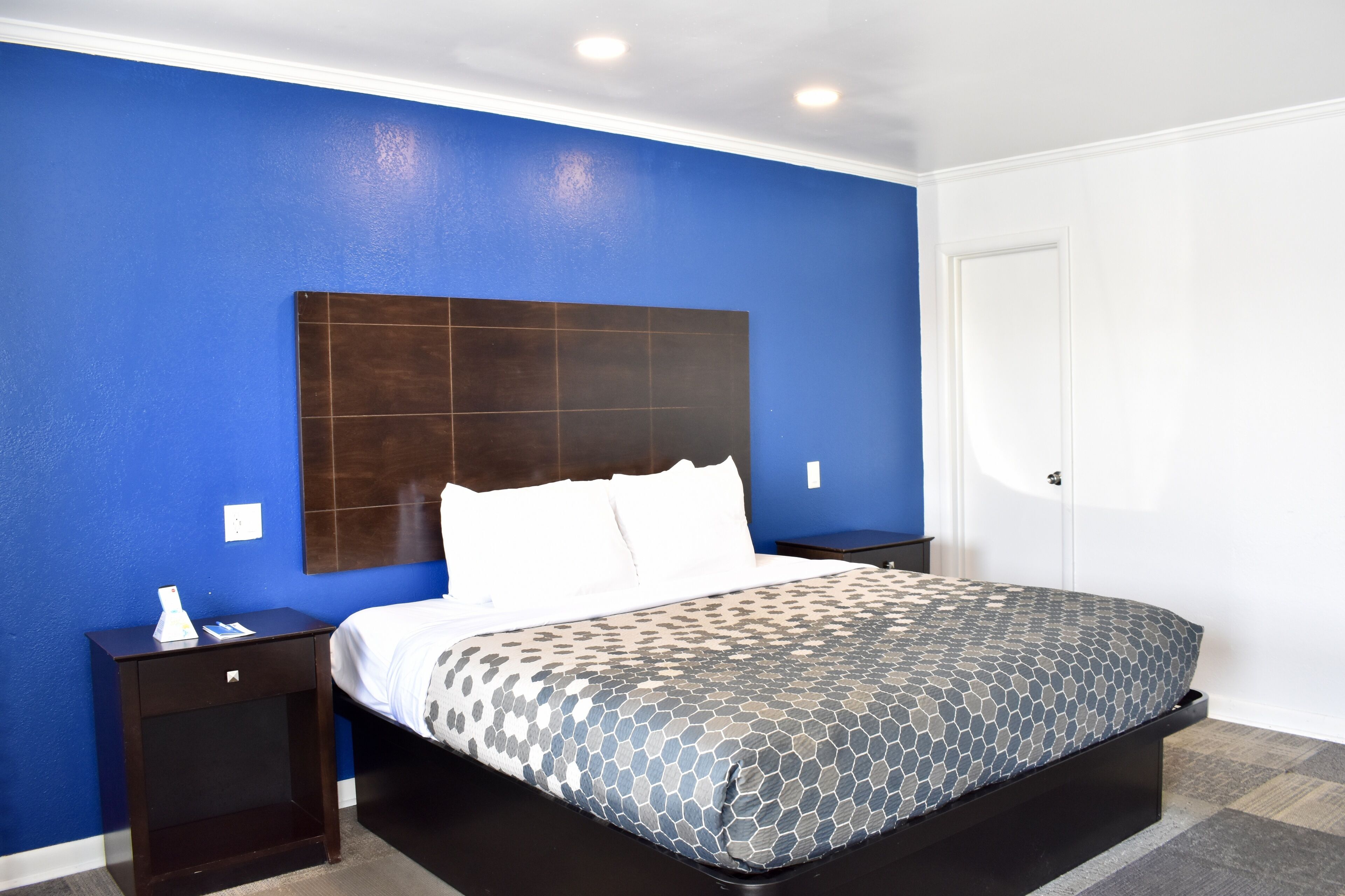 A hotel room with a blue accent wall, a large bed with a patterned bedspread, a dark headboard, and a nightstand with a phone and a small table.