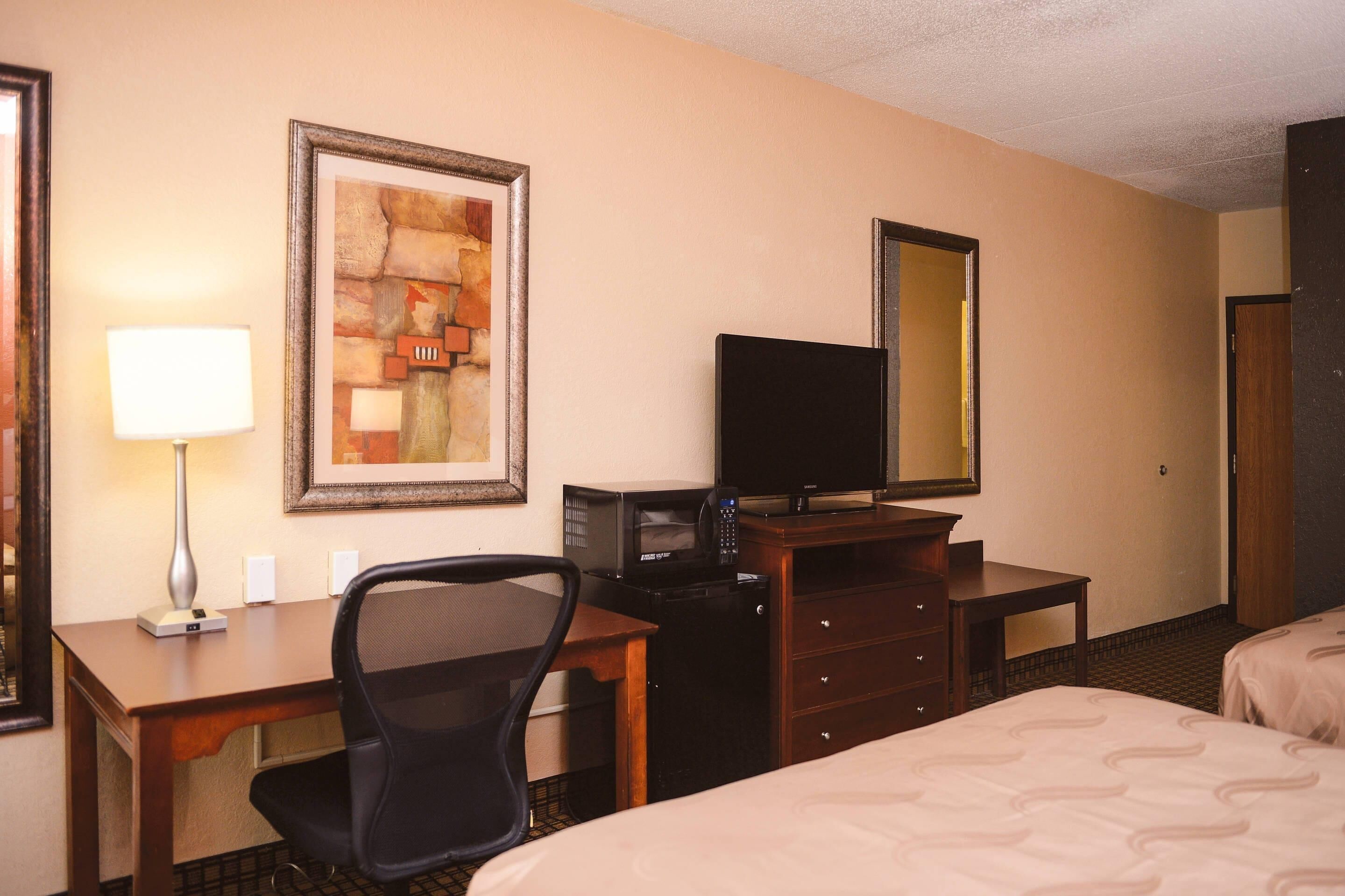 A hotel room with a bed, a desk with a chair, a television, a dresser, a framed artwork, and a mirror.