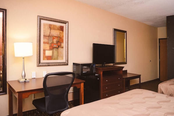 Habitación de hotel con cama, escritorio con silla, televisor, cómoda, obra de arte enmarco y espejo.