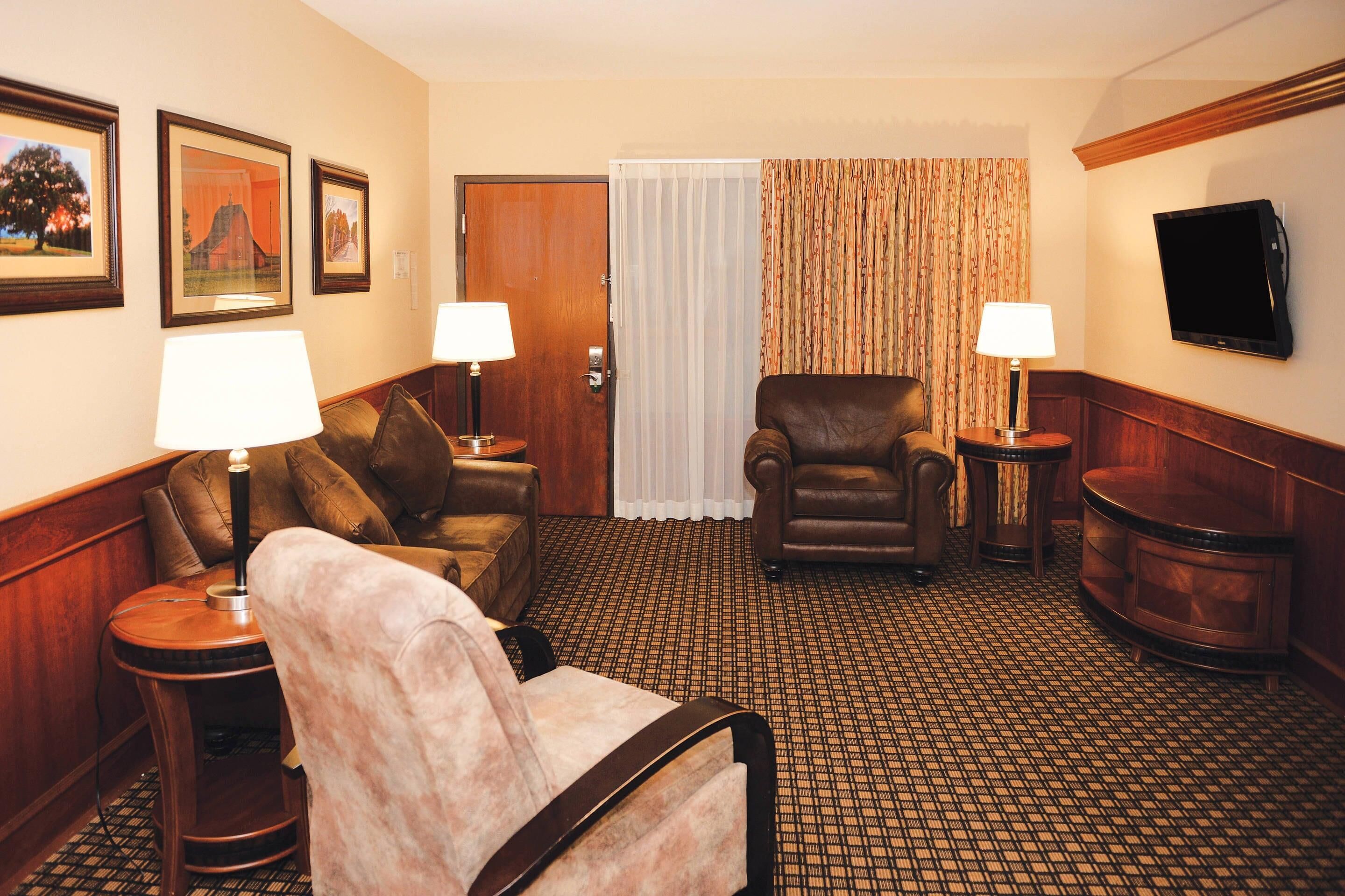 A hotel room with a sofa, armchair, and two side tables with lamps. There is a door with a curtain, a television mounted on the wall, and framed pictures on the wall.