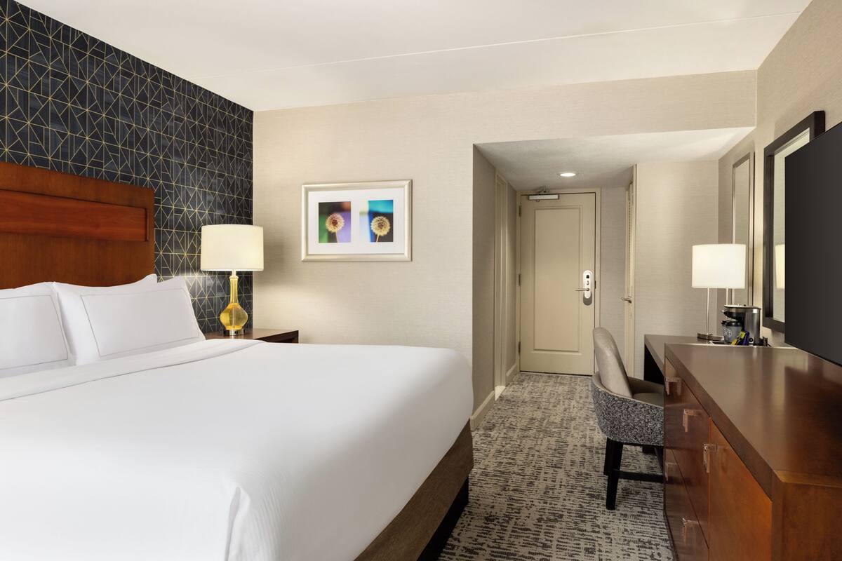 Ein Hotelzimmer mit einem großen Bett, einem Schreibtisch mit Stuhl, einem Fernseher und einem gerahmten Bild an der Wand.