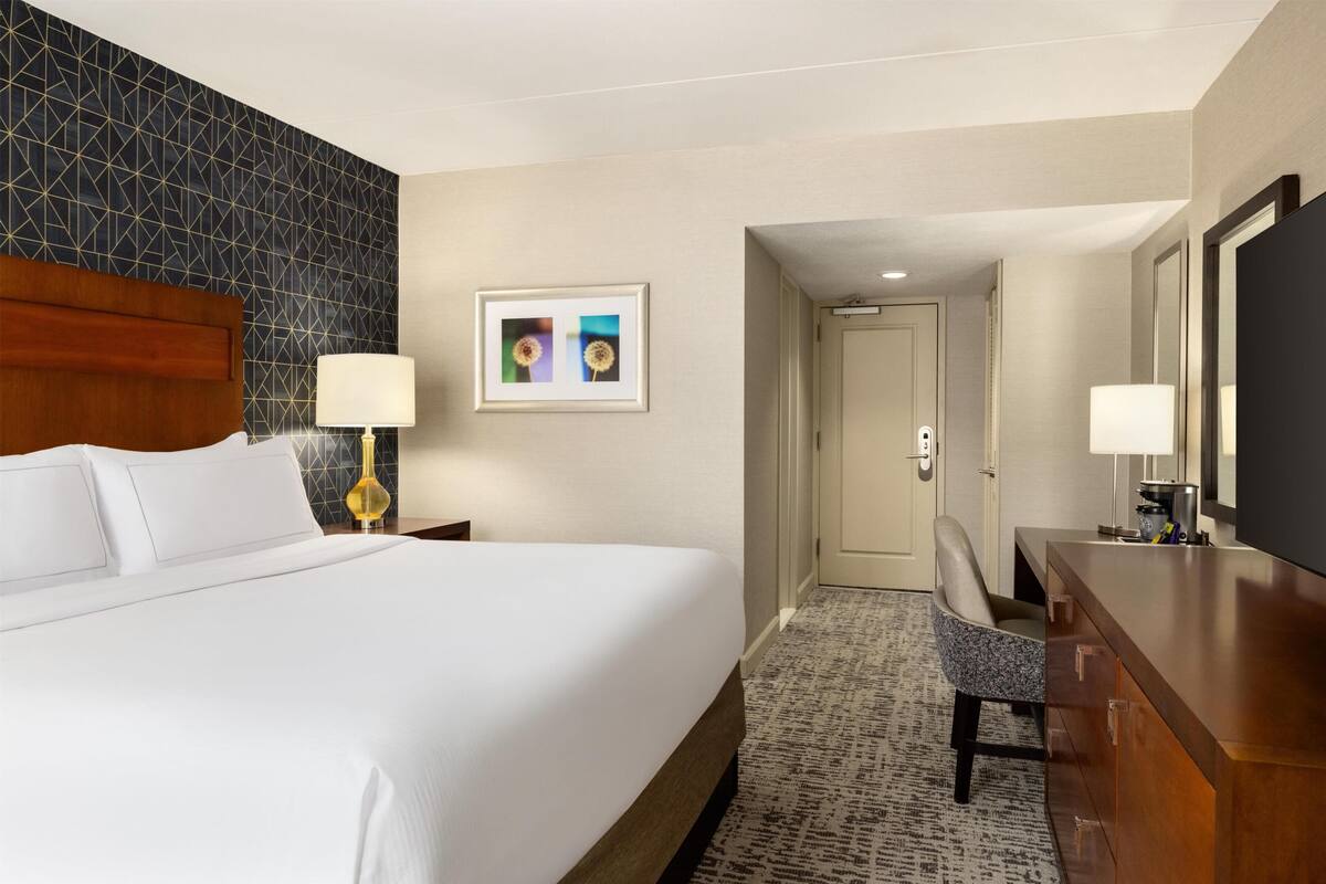 Ein Hotelzimmer mit einem großen Bett, einem Schreibtisch mit Stuhl, einer Lampe, einem gerahmten Bild an der Wand und einer Tür zu einem weiteren Raum.
