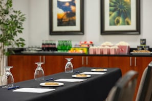 A conference table with plates of food, glasses, and pens, set against a backdrop of framed artwork and a buffet area.