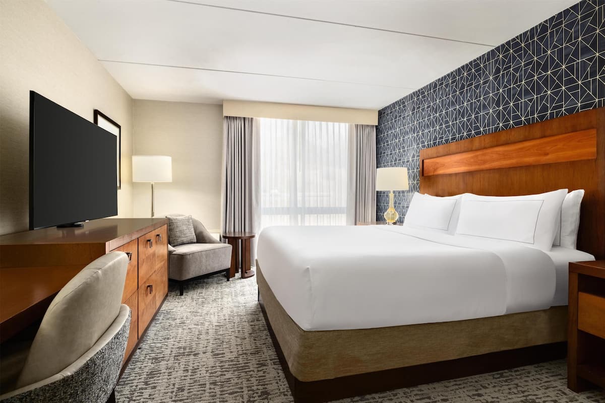 Ein Hotelzimmer mit einem Bett, einem Schreibtisch mit Fernseher, einem Sessel, einer Lampe und einem Fenster mit Vorhängen.
