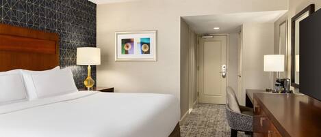 A hotel room with a large bed, a desk, a chair, a lamp, a framed picture, and a door leading to another room.