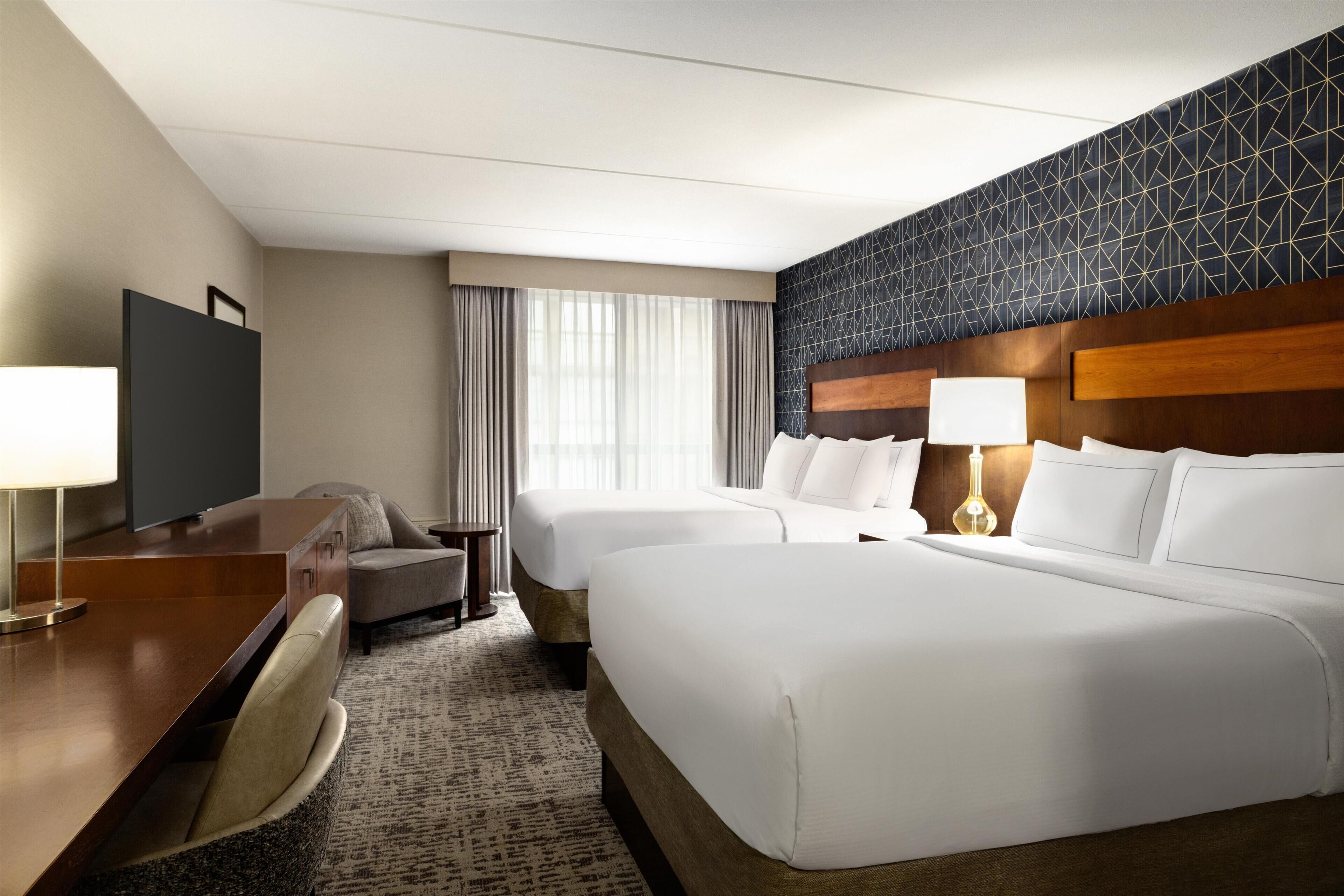 Ein Hotelzimmer mit einem großen Bett, einem Schreibtisch mit Stuhl, einem Fernseher und einem Fenster mit Vorhängen.